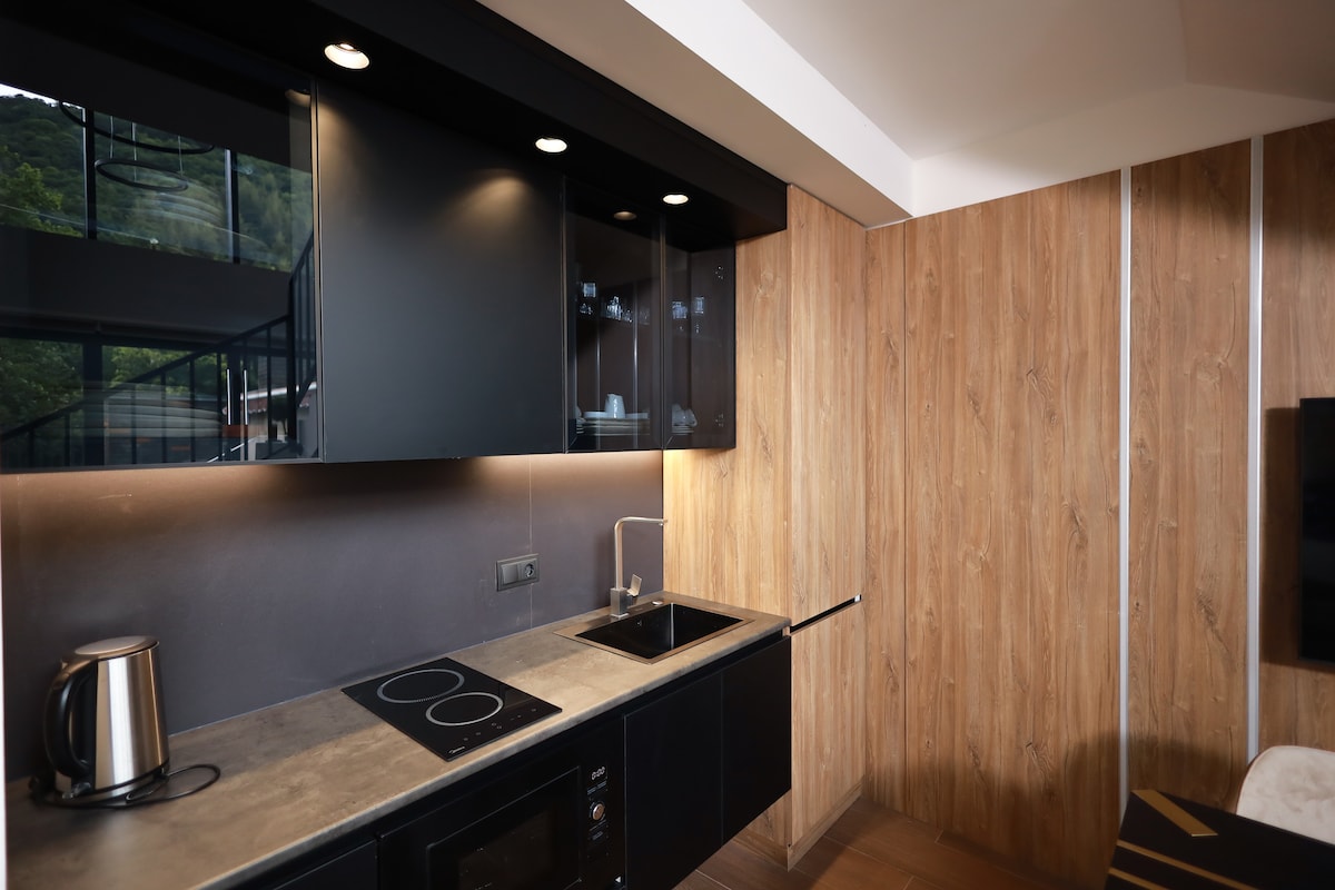 A well-equipped kitchenette features sleek black cabinets and modern appliances. A stovetop and microwave are integrated into the countertop, complemented by a kettle. Natural light filters through a nearby window, illuminating the textured wall's wood finish, creating a functional cooking space.