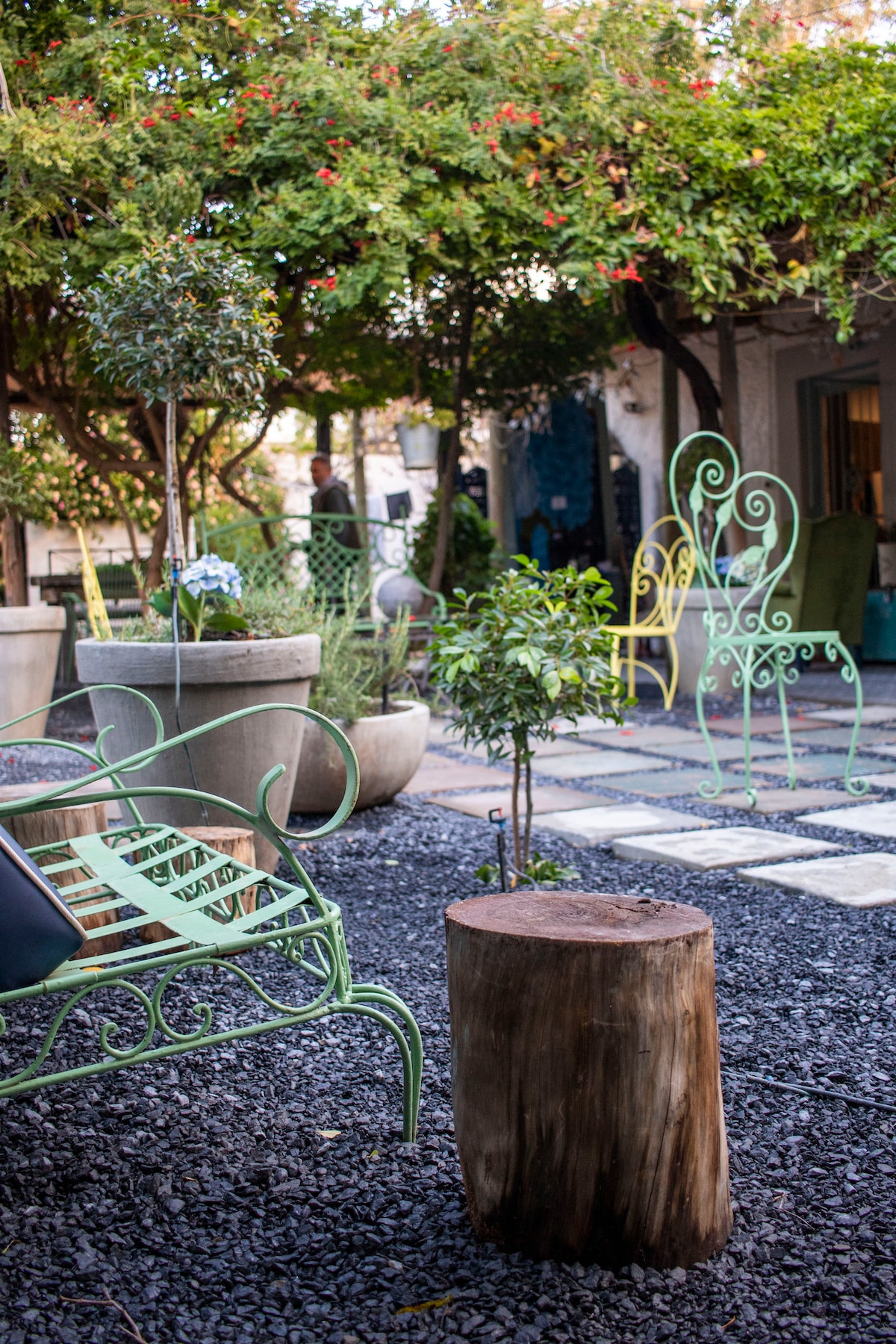 An outdoor seating area is depicted, featuring metallic chairs with decorative designs in shades of green. A wooden stool sits centrally on the gravel surface, surrounded by potted plants and lush greenery. The setting offers a serene and inviting space for relaxation.
