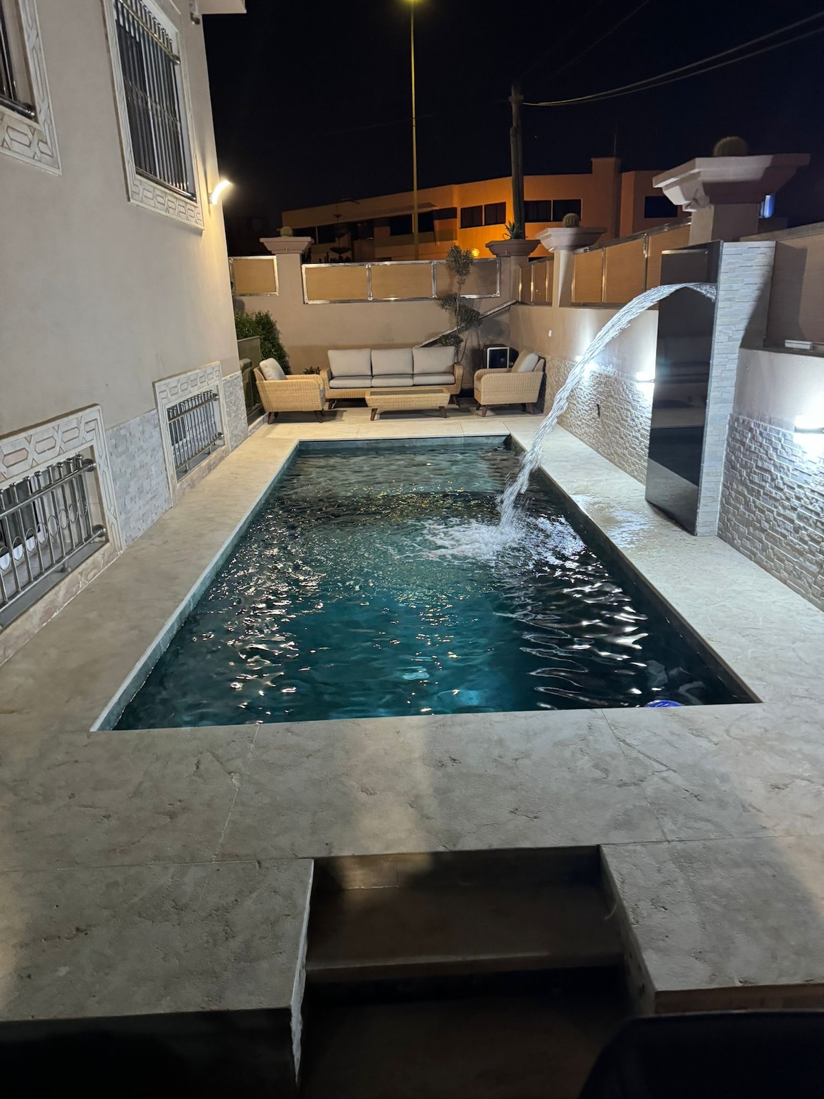 The private pool is illuminated under the night sky, featuring a gentle water feature. Adjacent to the pool, a seating area is furnished with comfortable lounge chairs, creating a relaxing outdoor space for enjoyment.