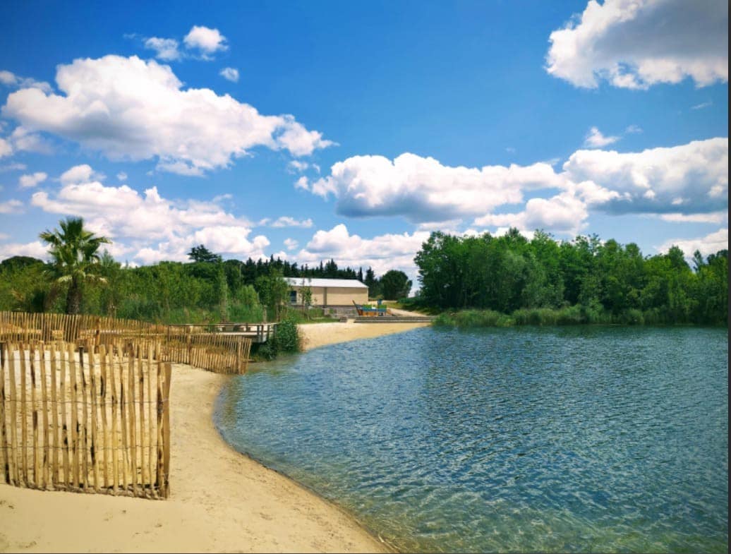 A sandy beach leads to calm waters, surrounded by lush greenery and wooden fencing. The scene is brightened by a partly cloudy sky, offering a serene outdoor space ideal for relaxation and enjoyment of nature.