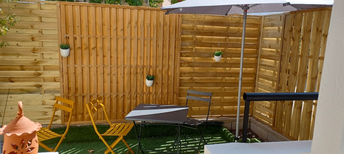 An outdoor space is enclosed by wooden fencing, featuring a small table and two chairs. A patio umbrella provides shade, while potted plants are mounted on the fence. The area is adorned with artificial grass, enhancing the serene atmosphere.