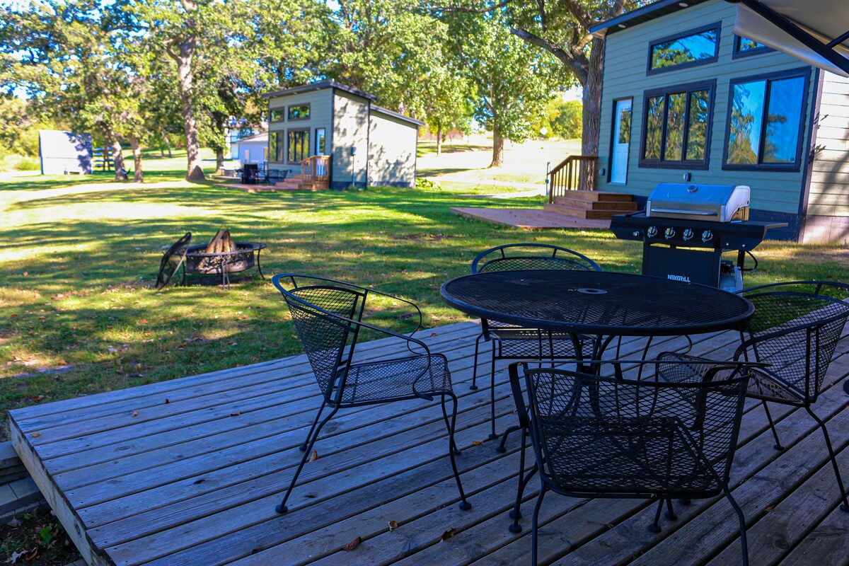 An outdoor seating area is presented on a wooden deck, featuring a round table and several black metal chairs. A propane grill is positioned nearby, while a fire pit sits in the grassy area, surrounded by trees and other camper cabins.