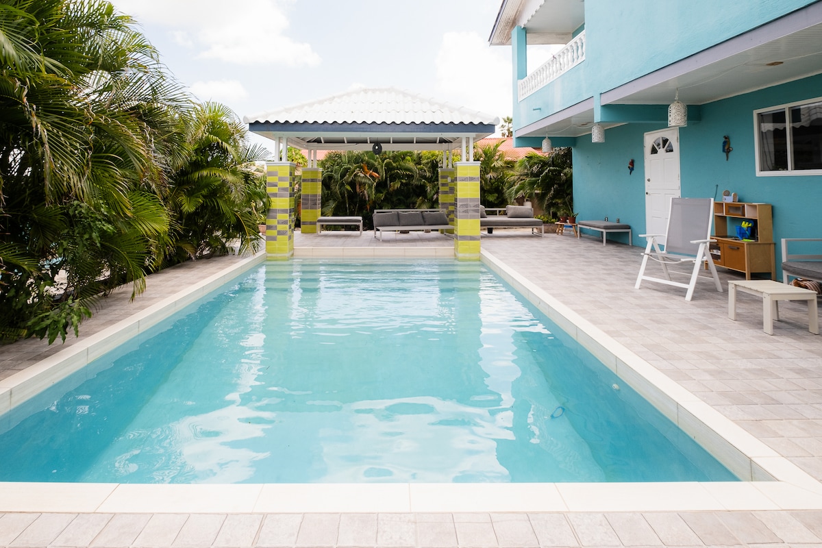 A shared swimming pool features crystal-clear water, surrounded by tiled decking and tropical foliage. Comfortable lounge chairs offer a space for relaxation, and a gazebo provides shaded seating nearby. A vibrant blue building stands in the background, completing the inviting outdoor setting.