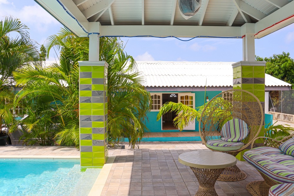 An outdoor area features a swimming pool surrounded by tropical palm trees. Colorful accent walls are displayed on the adjacent building, while a shaded seating area is furnished with a round table and two hanging chairs, inviting relaxation in a cheerful atmosphere.
