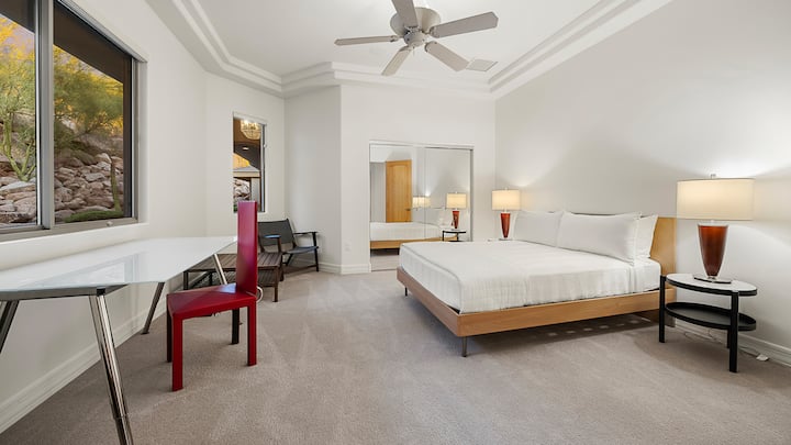 Bright and modern bedroom with a desk, tray ceiling, and serene views of trees and natural rock formations.