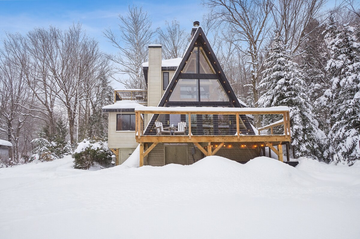 A rustic chalet with a distinctive A-frame design is surrounded by a snowy landscape, showcasing a spacious wooden deck with built-in seating. Large glass windows provide a view of the winter scenery, allowing natural light to fill the interior. Snow blankets the ground, enhancing the serene environment.