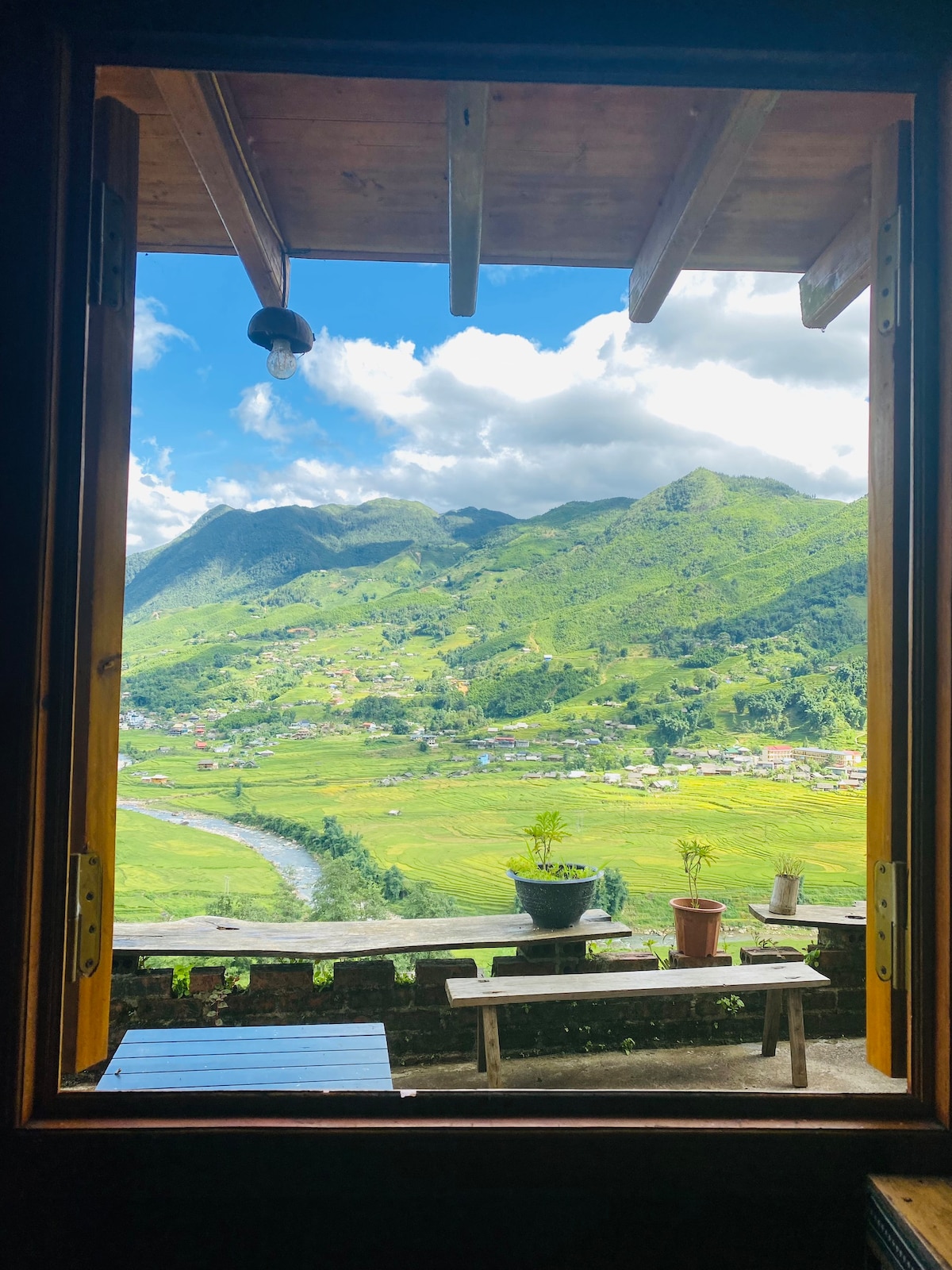 A large window frames a scenic view of the Muong Hoa valley, featuring lush green mountains and terraced fields. A wooden bench and potted plants are visible on the balcony, enhancing the connection to nature. Soft clouds float in a bright blue sky.