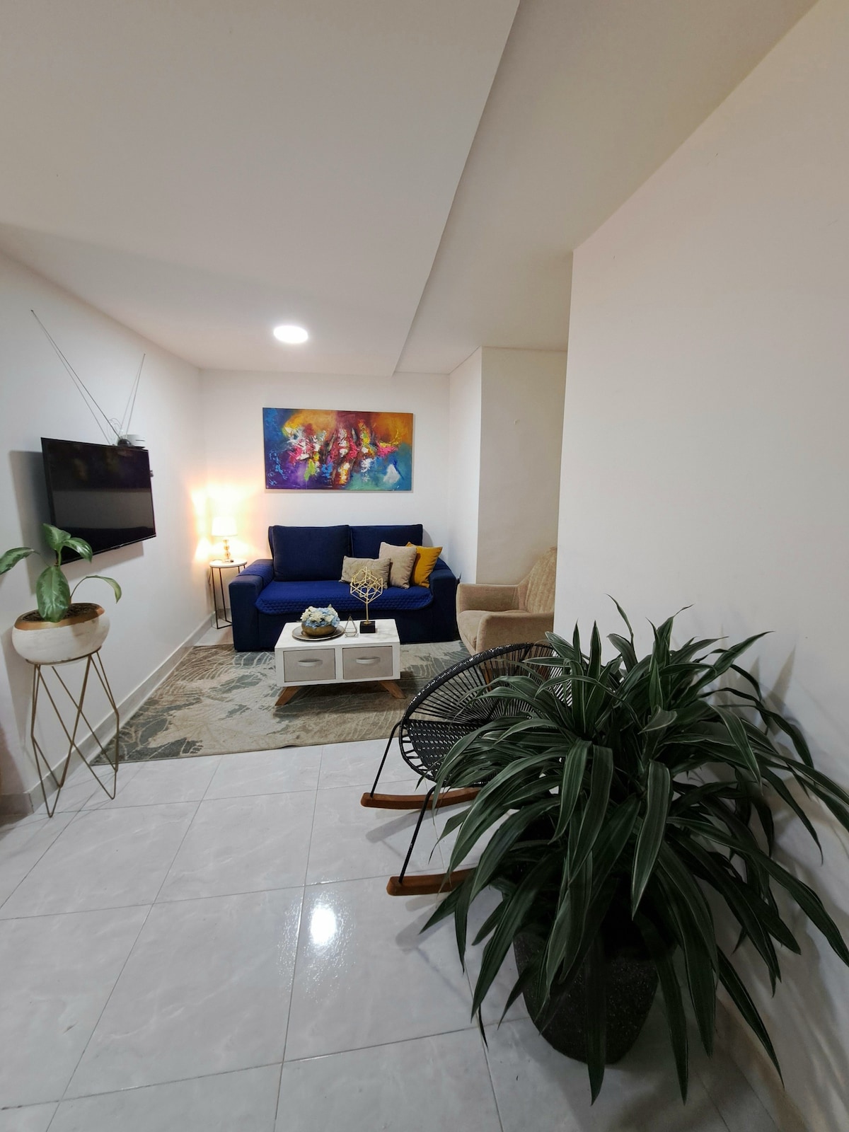 A cozy living area features a blue sofa adorned with colorful cushions, complemented by a stylish accent chair. A coffee table holds decorative items, while a potted plant adds natural touches. Soft lighting creates a welcoming environment, with a vibrant piece of artwork enhancing the wall.