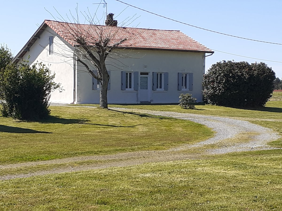 A charming house with a white facade and a red-tiled roof is surrounded by greenery and well-maintained grass. A gravel path leads to the front door, while trees and shrubs are positioned thoughtfully on the property, adding to the peaceful setting.