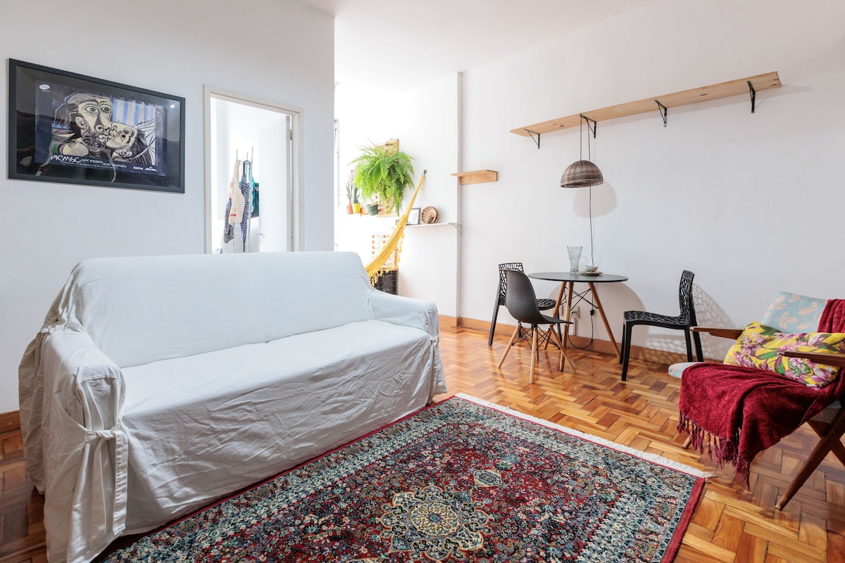 A bright living area features a white slipcovered sofa, a round dining table with two chairs, and an area rug with intricate patterns. A shelf holds decorative items, and natural light enters through the doorway, contributing to an inviting ambiance.