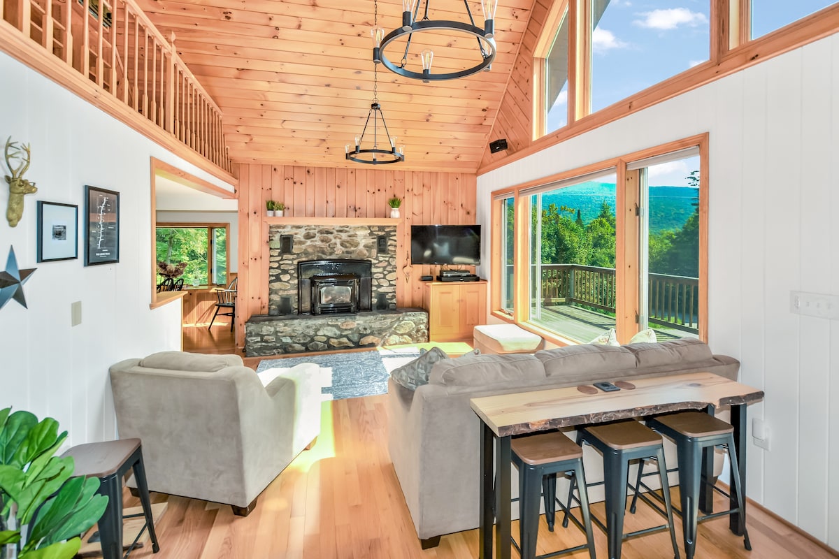 A spacious living area features natural wood paneling and a stone fireplace as the focal point. Large windows allow for ample natural light and views of the mountains. Comfortable seating arrangements include a pair of sofas and a dining table with stools, creating a welcoming gathering space.