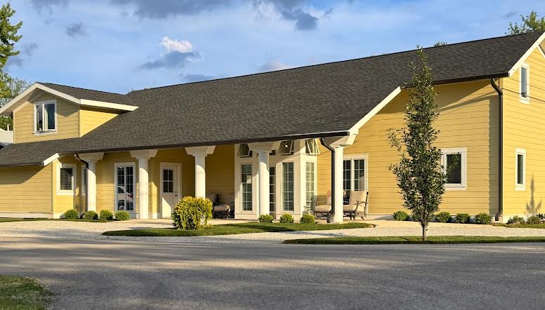 The exterior of a yellow, modern building is shown, featuring a sturdy roof and a wraparound porch with multiple seating options. Lush greenery borders the pathway, and tall windows allow natural light to illuminate the interior spaces.