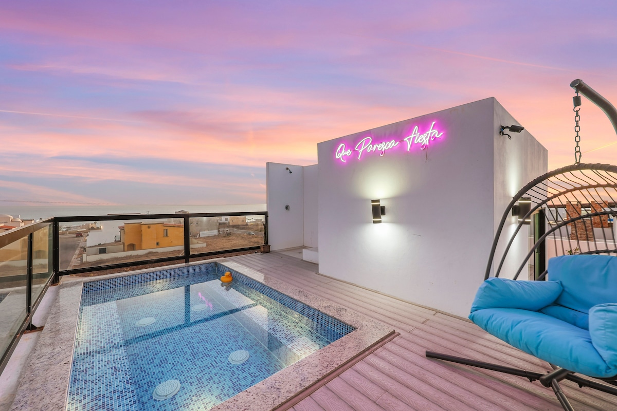 A rooftop area features a small, inviting pool surrounded by a wooden deck. A stylish hanging chair is positioned nearby. A neon sign adds a pop of color against the serene backdrop of a pastel sunset sky.
