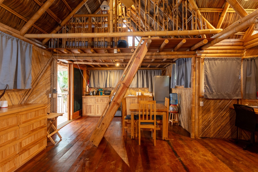 The interior of the bamboo bungalow features an open-concept design with wooden floors and walls. A dining table with chairs occupies the space, accompanied by a kitchenette equipped with essential appliances. Natural light filters in through windows, enhancing the warm tones of the bamboo.