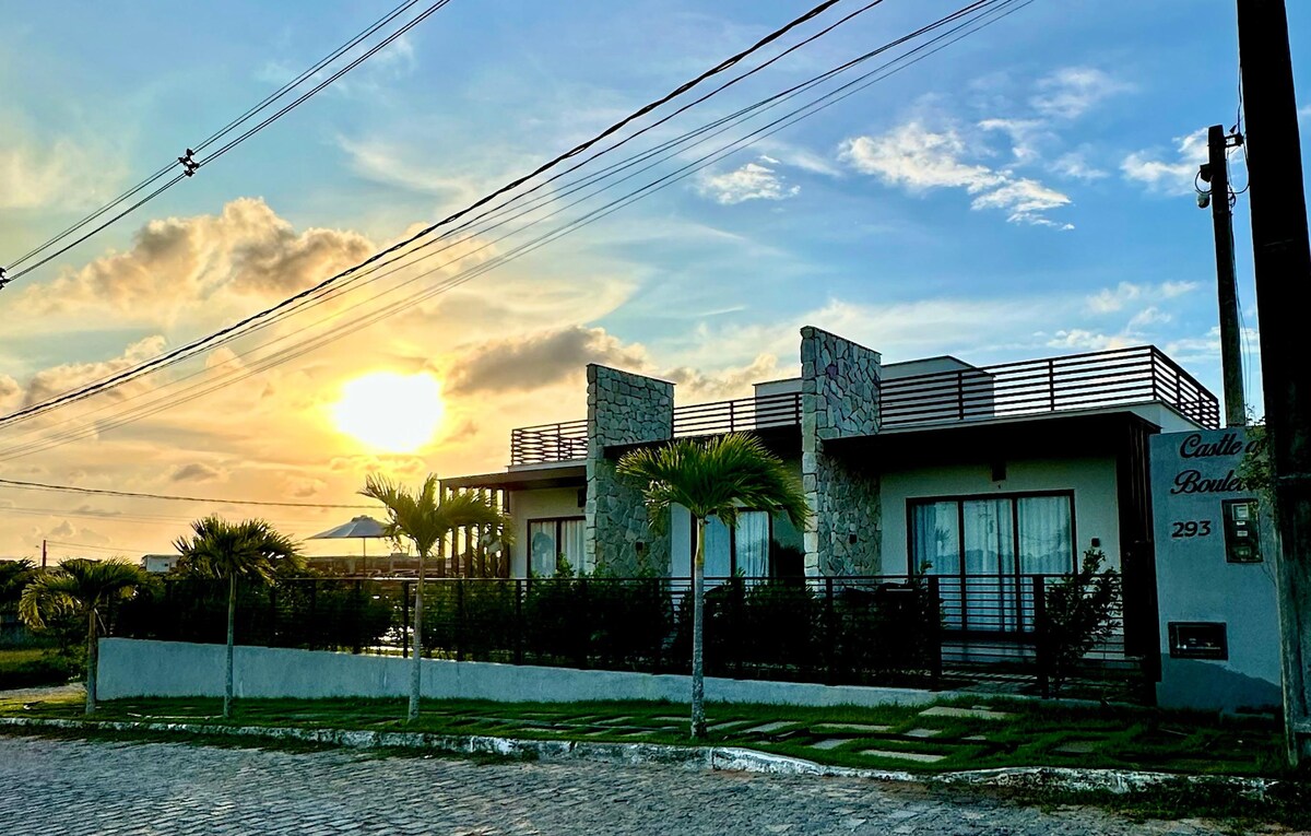 A modern chalé is seen against a sunset, with a stone façade and wide glass doors. Lush greenery surrounds the property, complemented by palm trees. The rooftop area is visible, allowing for outdoor enjoyment and scenic views.