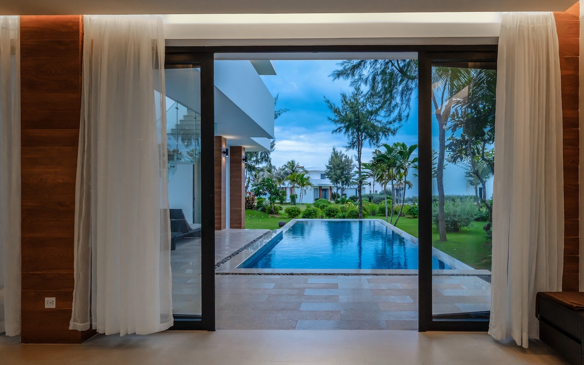 Large glass doors provide a view of a private pool surrounded by lush greenery. The tranquil outdoor space features palm trees and well-maintained grass, contributing to a serene atmosphere. Soft, flowing curtains frame the view, complementing the modern interior.
