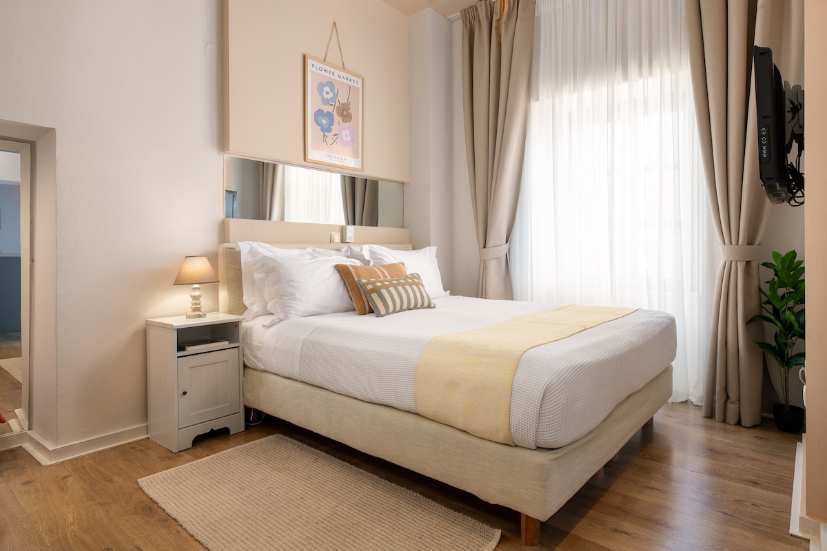 A bright and airy bedroom features a spacious bed covered with a white duvet and a light yellow throw. A bedside table holds a lamp, and large windows with sheer curtains allow natural light to fill the room. A decorative wall art piece hangs above the bed.