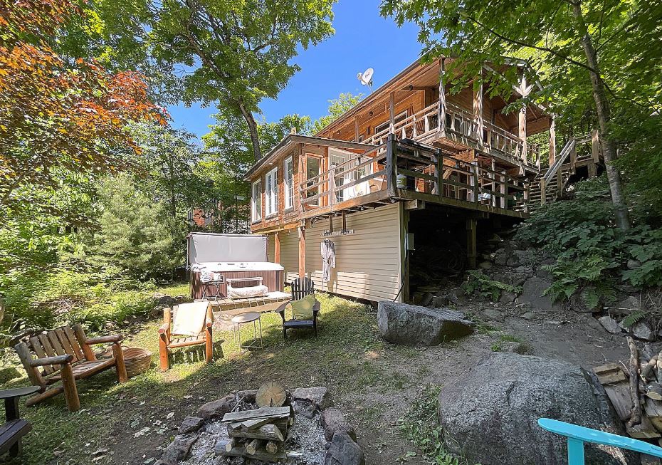 The rustic chalet is nestled among greenery, featuring a spacious terrace with seating and a hot tub. A fire pit sits on a stone path leading to the home, which is elevated and surrounded by trees. Large windows offer a view of the serene environment.
