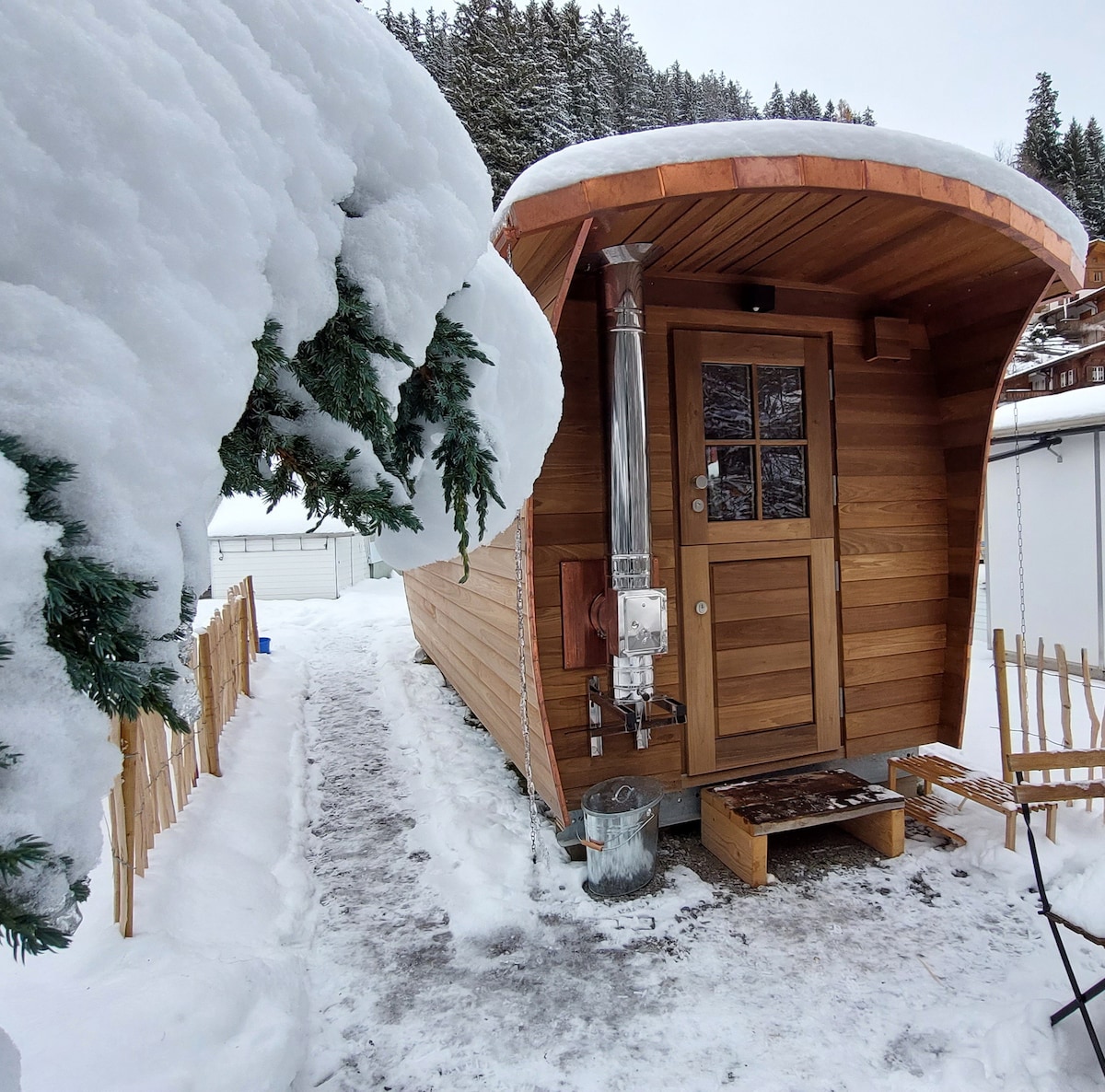 The tiny wooden house features a rounded roof and is surrounded by snow-covered trees. A pathway is visible leading to the front door, with a wood stove located beside the entrance. Soft snow blankets the ground and roofing, creating a peaceful winter scene.