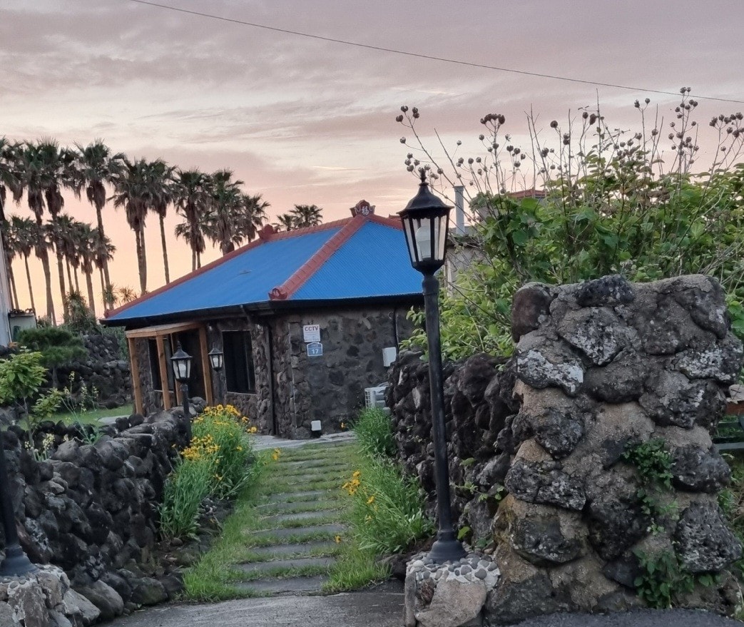 A stone pathway leads to a charming house with a blue roof, framed by lush greenery and palm trees. A stone wall surrounds the property, while a lantern guides the way. The soft light of dusk enhances the welcoming facade.