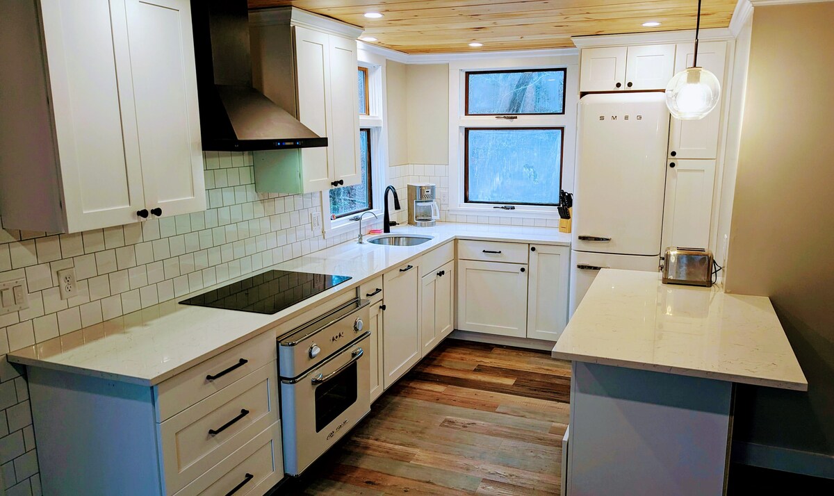 A modern kitchen is displayed with white cabinetry and a light wood ceiling. An induction cooktop and a BigChill oven are visible, accompanied by a SMEG refrigerator. Natural light enters through a window above the sink, illuminating the spacious countertops and tile backsplash.