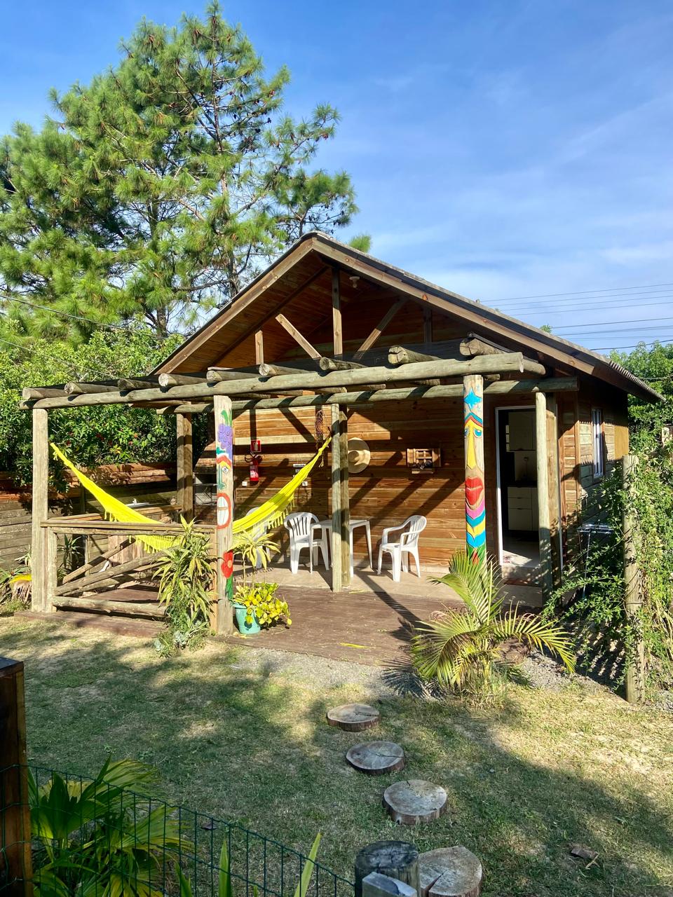 A charming wooden cabin is set among greenery, featuring a porch supported by rustic beams. Two simple white chairs and colorful hammocks invite relaxation. A pathway of stepping stones leads to the entrance, surrounded by landscaped grass and plants.