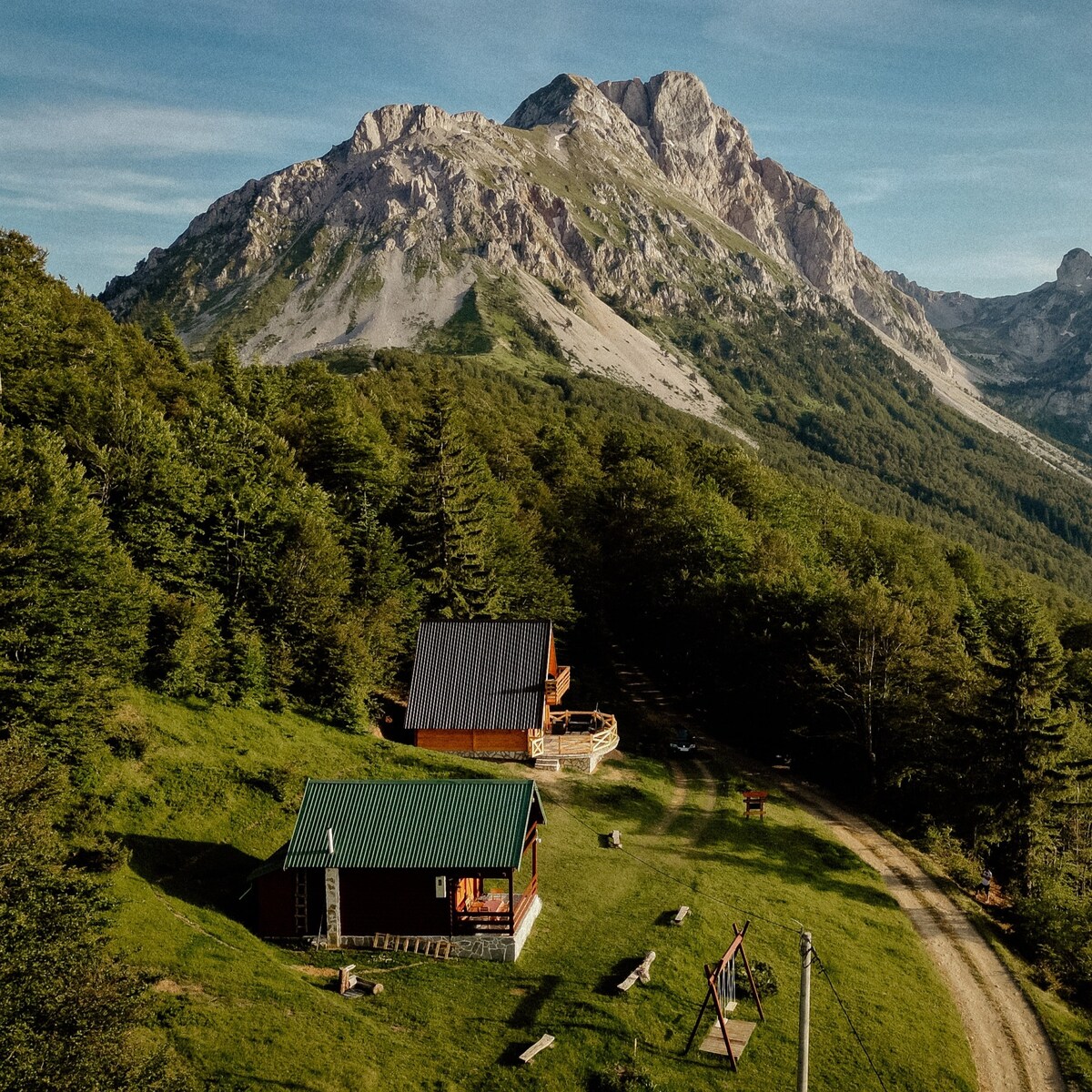 A serene view showcases two rustic cabins nestled among lush greenery, with a backdrop of towering mountains. A winding dirt path leads through the landscape, while logs are placed around the cabins, inviting outdoor enjoyment in a peaceful, natural setting.