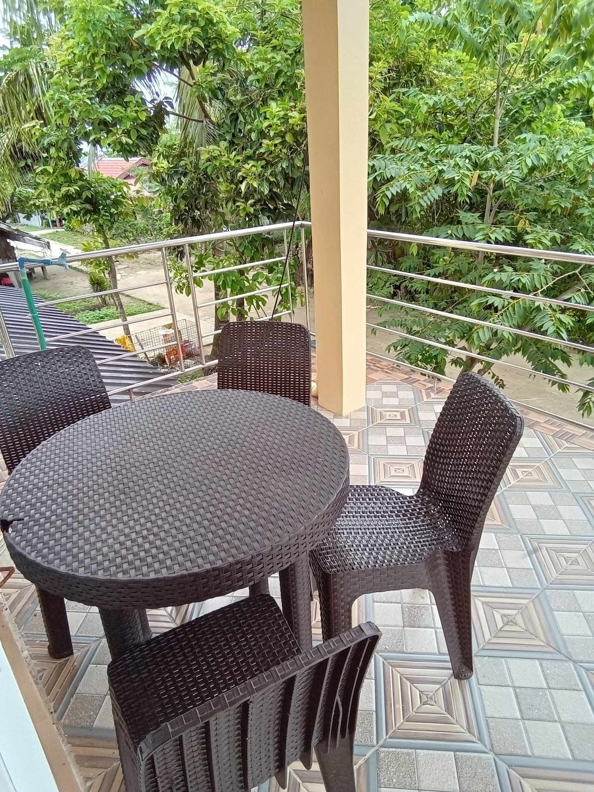 An outdoor seating area is featured on a balcony, showcasing a round table surrounded by four woven chairs. Lush greenery borders the space, providing a natural ambiance, while the tiled floor adds a touch of character to the setting.