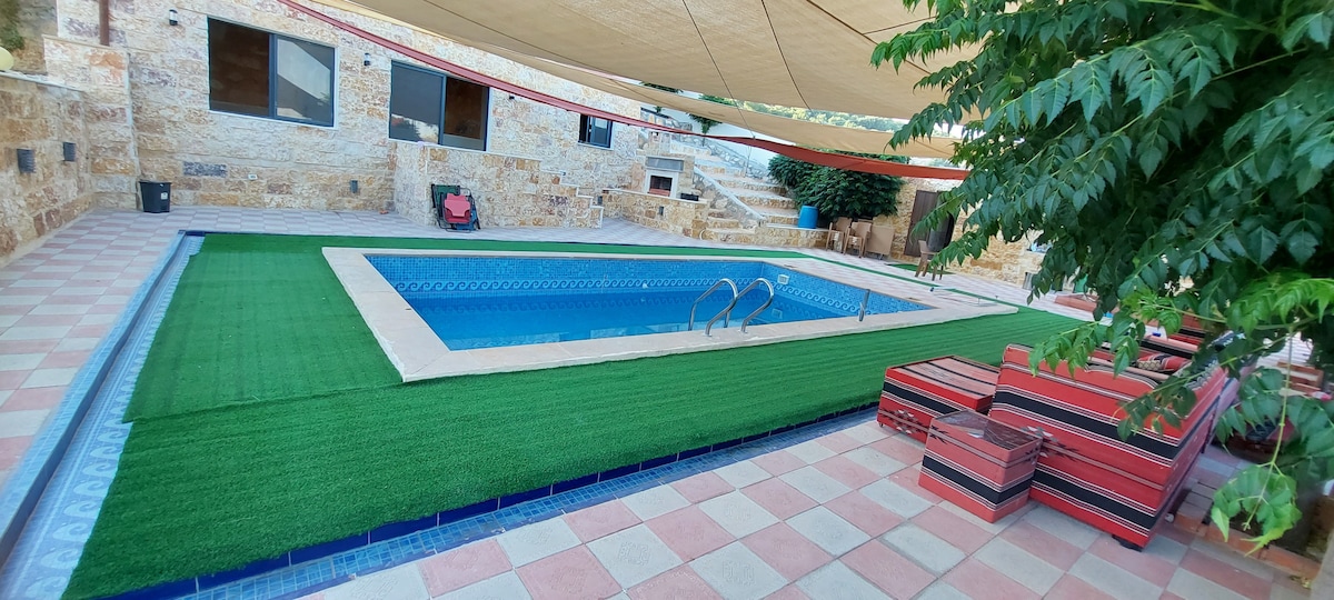 A swimming pool is surrounded by a spacious patio area, featuring artificial grass and shade from canopy structures. Colorful lounge seating provides an area for relaxation, while natural stone walls frame the scene, enhancing the outdoor ambiance.