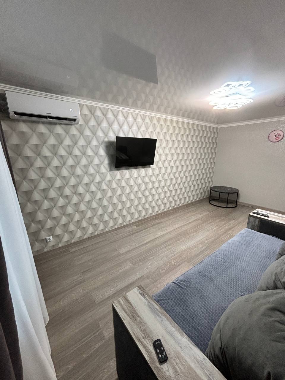 A modern living area features textured wall panels and a sleek entertainment setup with a wall-mounted television. Natural light enters through a nearby window, enhancing the space. A cozy sofa is accentuated by minimalist decor, offering a calm and inviting ambiance.