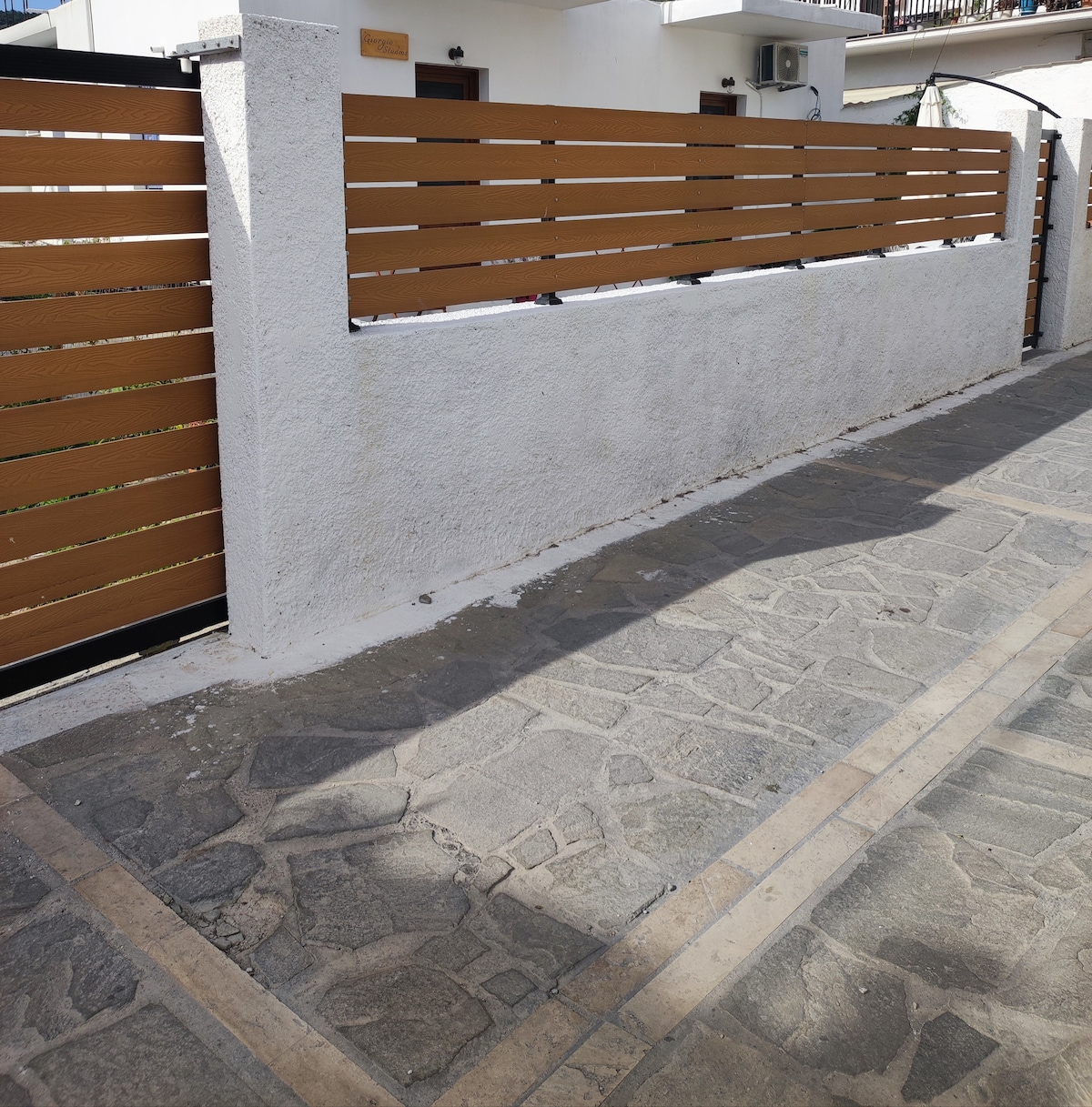 A pathway is depicted, featuring a textured stone surface and bordered by a modern wooden fence. The wall is painted white, contrasting with the wooden slats. Natural light casts soft shadows, highlighting the clean lines of the enclosure, which provides a sense of privacy.