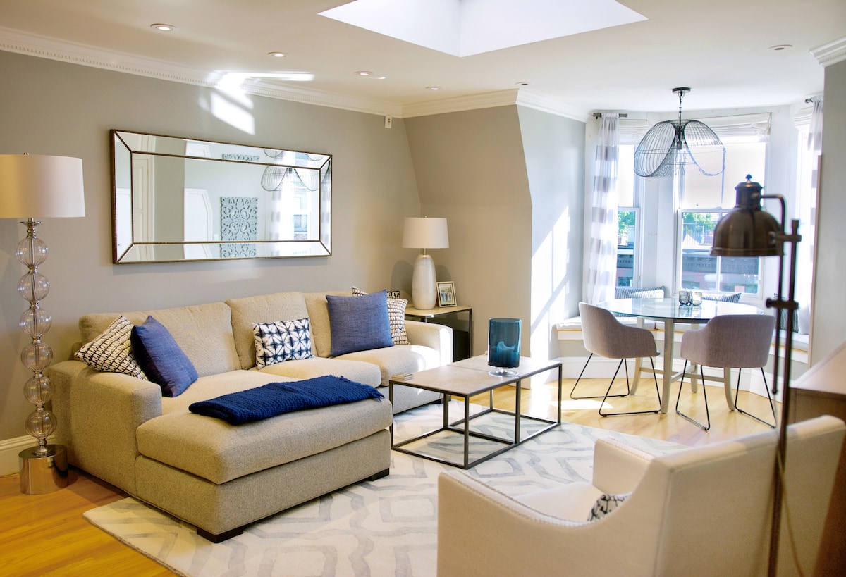 A bright living area features a spacious sectional sofa with decorative pillows, complemented by a coffee table. A dining table with modern chairs sits nearby, illuminated by ample natural light through large windows. Elegant decor, including a mirror and contemporary light fixtures, enhances the inviting atmosphere.
