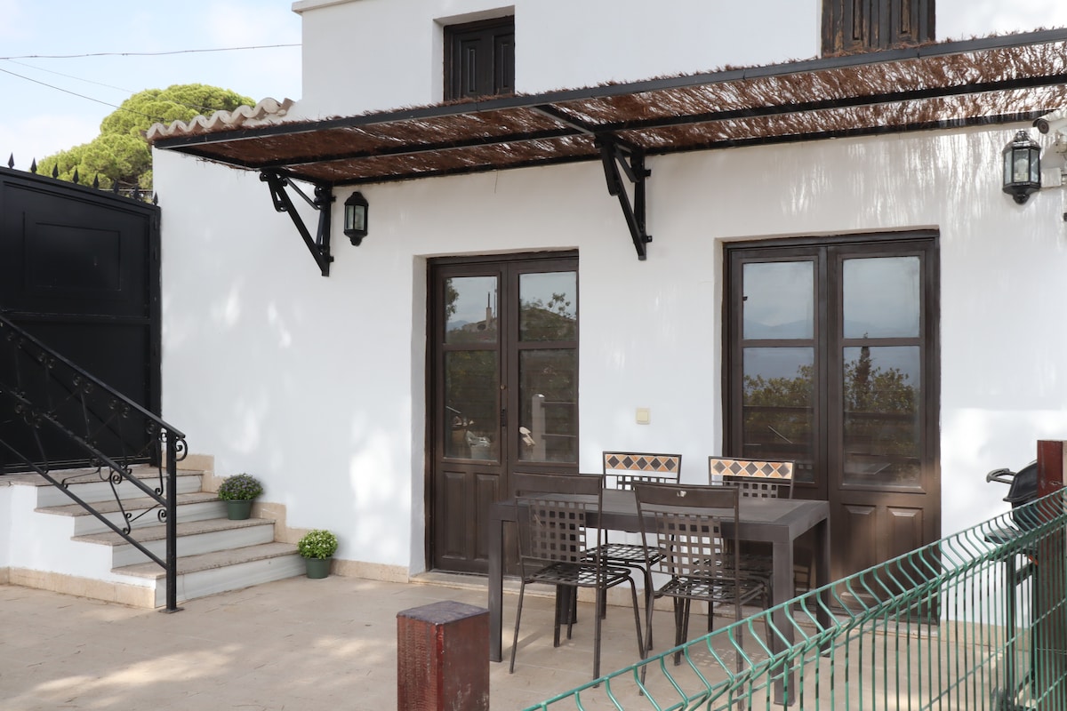 A covered outdoor dining area features a table with four chairs, set against a backdrop of a whitewashed building. Stairs lead to the entrance, and potted plants are placed near the base, adding a touch of greenery.