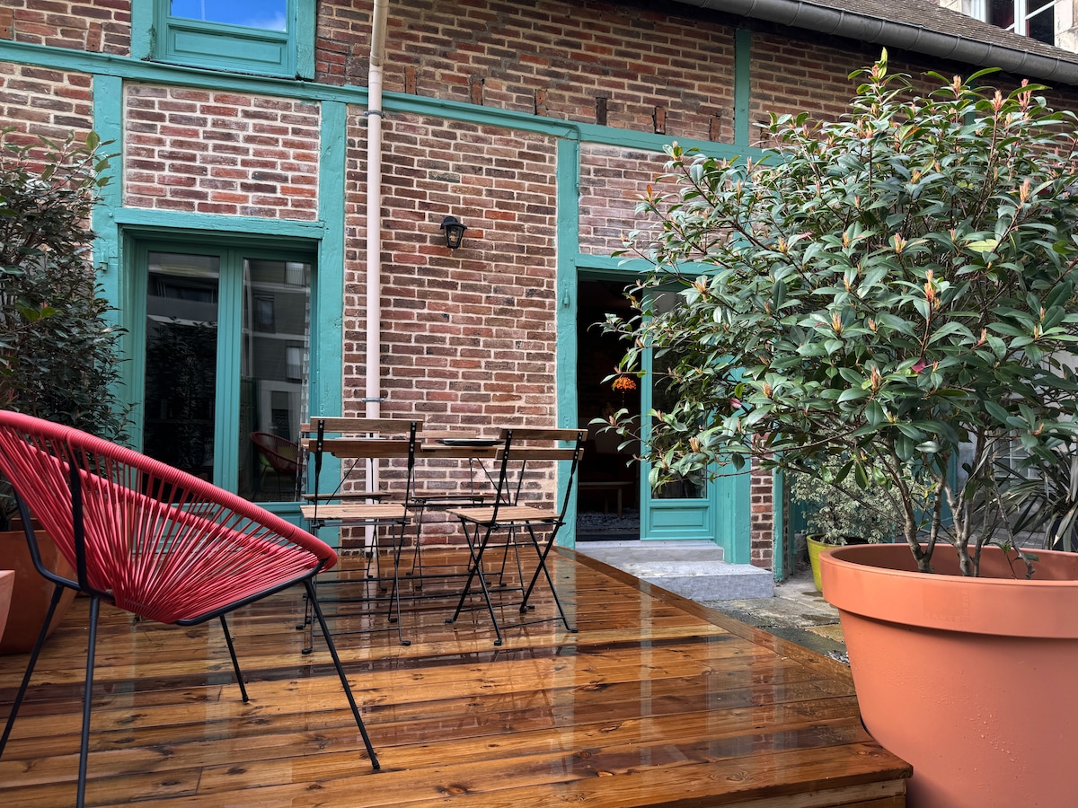 A wooden terrace features a round table surrounded by four black folding chairs. Near the seating area, a large potted plant adds greenery. The brick façade is highlighted by teal accents, creating a welcoming outdoor space.