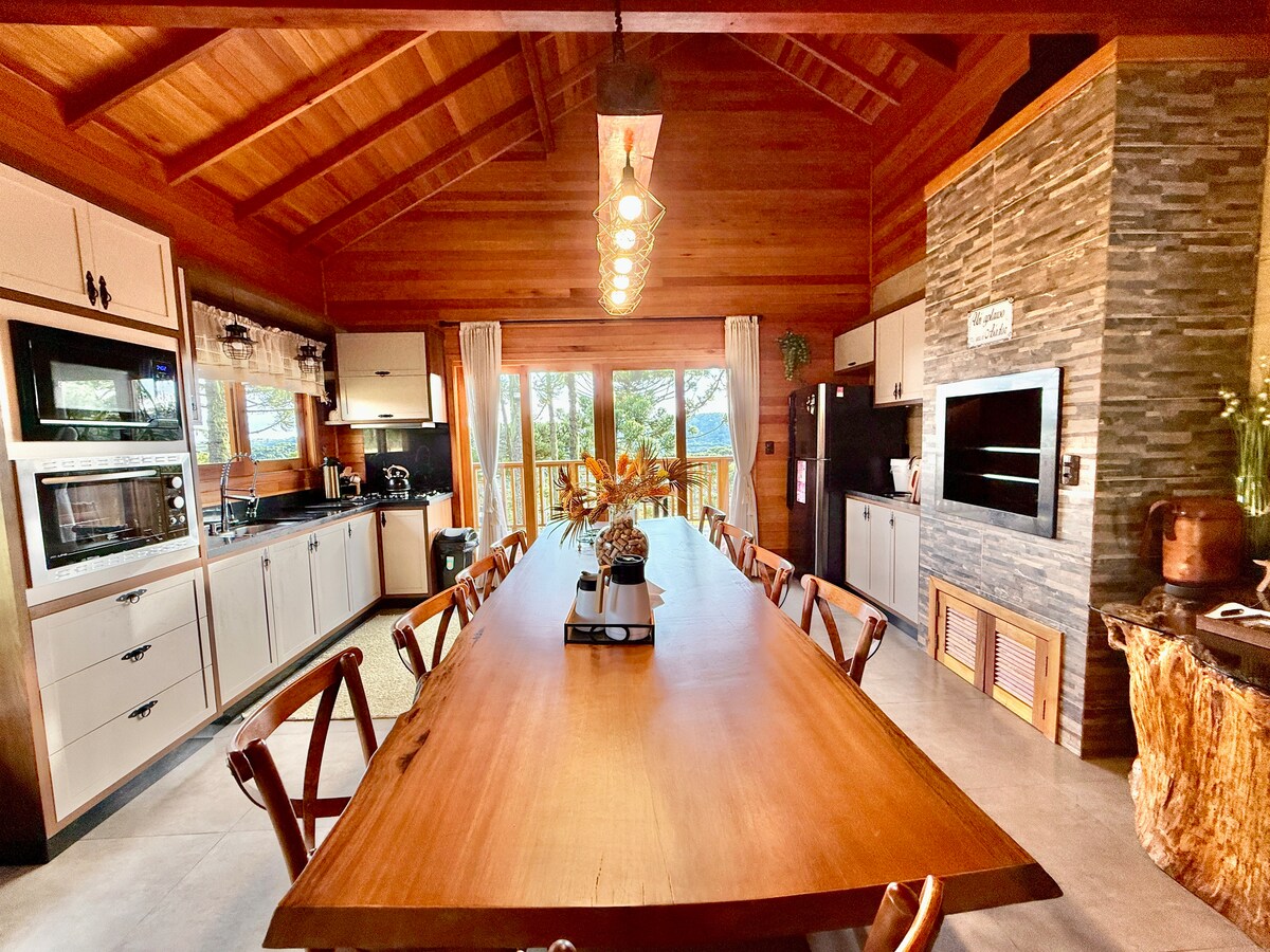 A spacious kitchen and dining area features a large wooden table surrounded by rustic chairs, complemented by large windows that provide natural light and views of the outdoor greenery. Modern appliances and wooden accents enhance the inviting atmosphere.