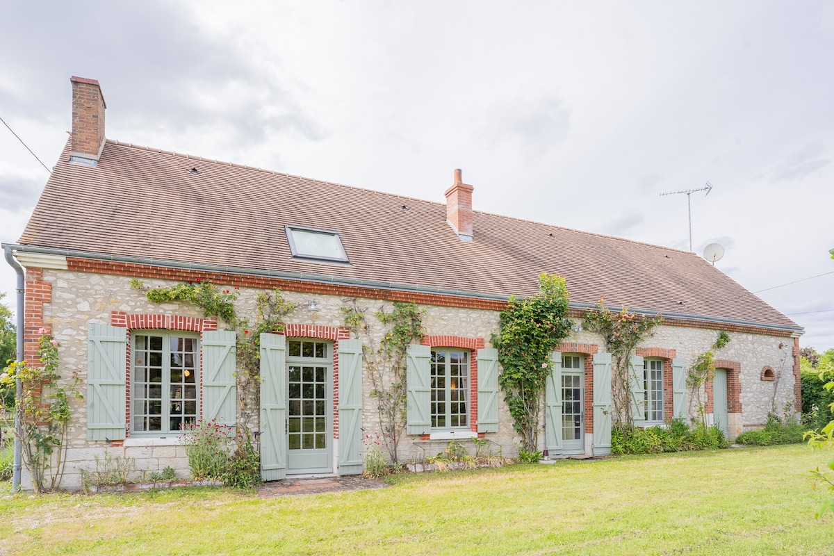 A charming single-story house is set in a lush green landscape, featuring brick accents and decorative window shutters. Climbing vines add greenery to the exterior, while large windows invite natural light into the interior. A peaceful environment is suggested by the well-maintained lawn.