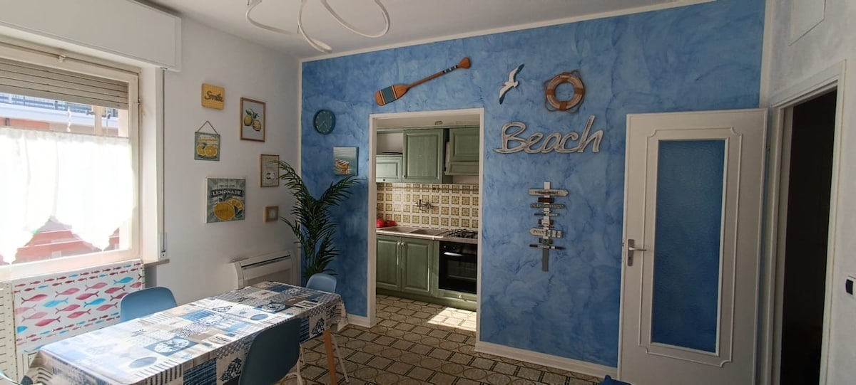 A light-filled dining area showcases a decorative blue wall adorned with beach-themed accents. A patterned table is surrounded by four blue chairs. The kitchen, featuring sea-inspired tiles and modern appliances, is visible through an open doorway.
