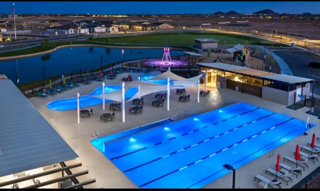 A community pool area is illuminated at night, featuring a lap pool and leisure pools surrounded by lounge areas. Umbrellas provide shade over tables, while lights highlight the water's vibrant blue. A nearby playground structure is visible, adding to the inviting outdoor space.