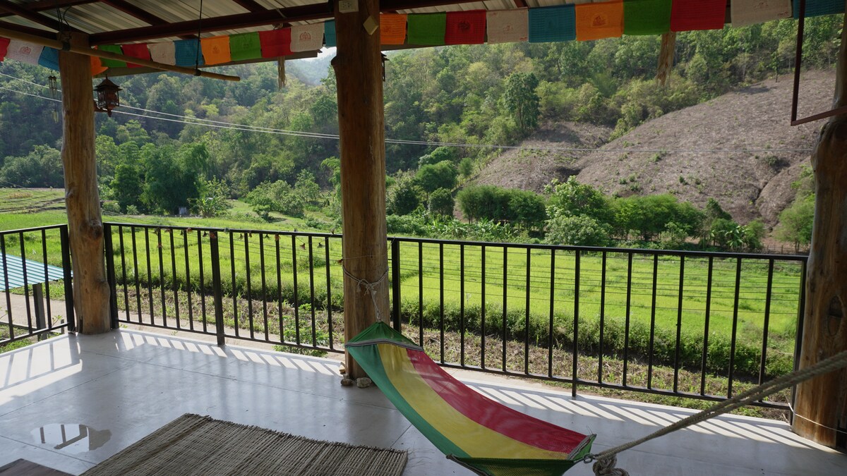 A spacious deck is featured, showcasing a colorful hammock and a table for four. Lush rice fields and mountains are visible in the background, framed by a wooden railing and colorful prayer flags hanging above.