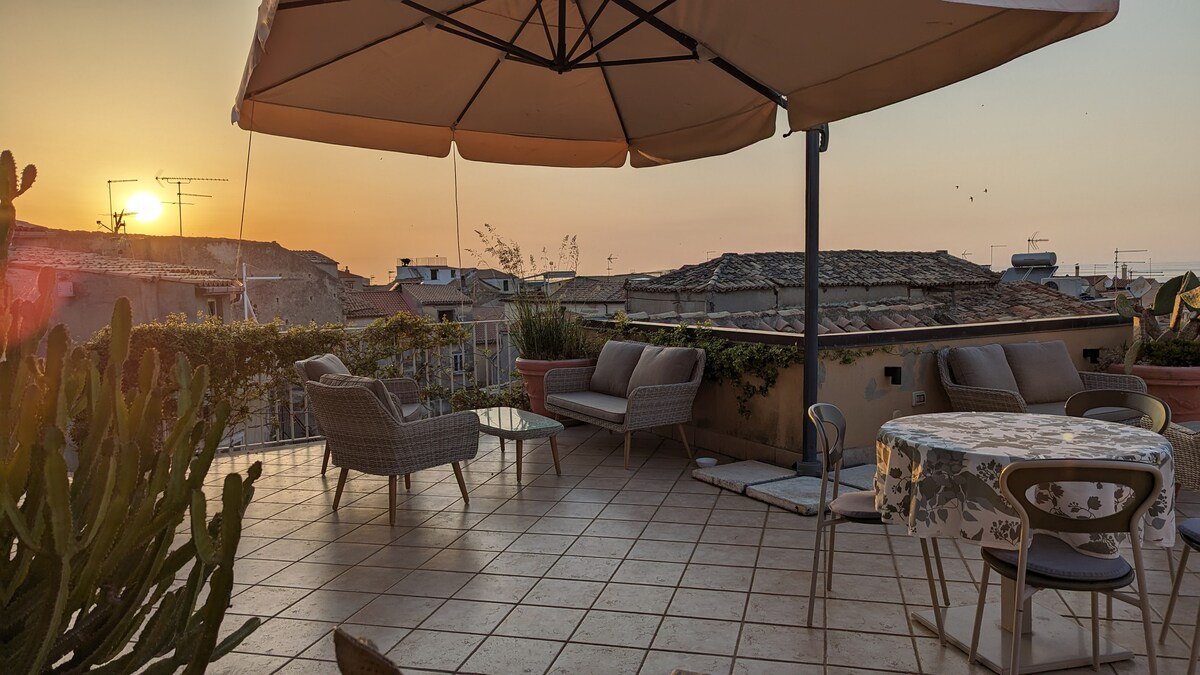 A spacious terrace showcases comfortable seating arrangements, including a round table and several lounge chairs. The area is surrounded by plants and features an umbrella for shade. A sunset view fills the background, casting warm light over the tiled floor.