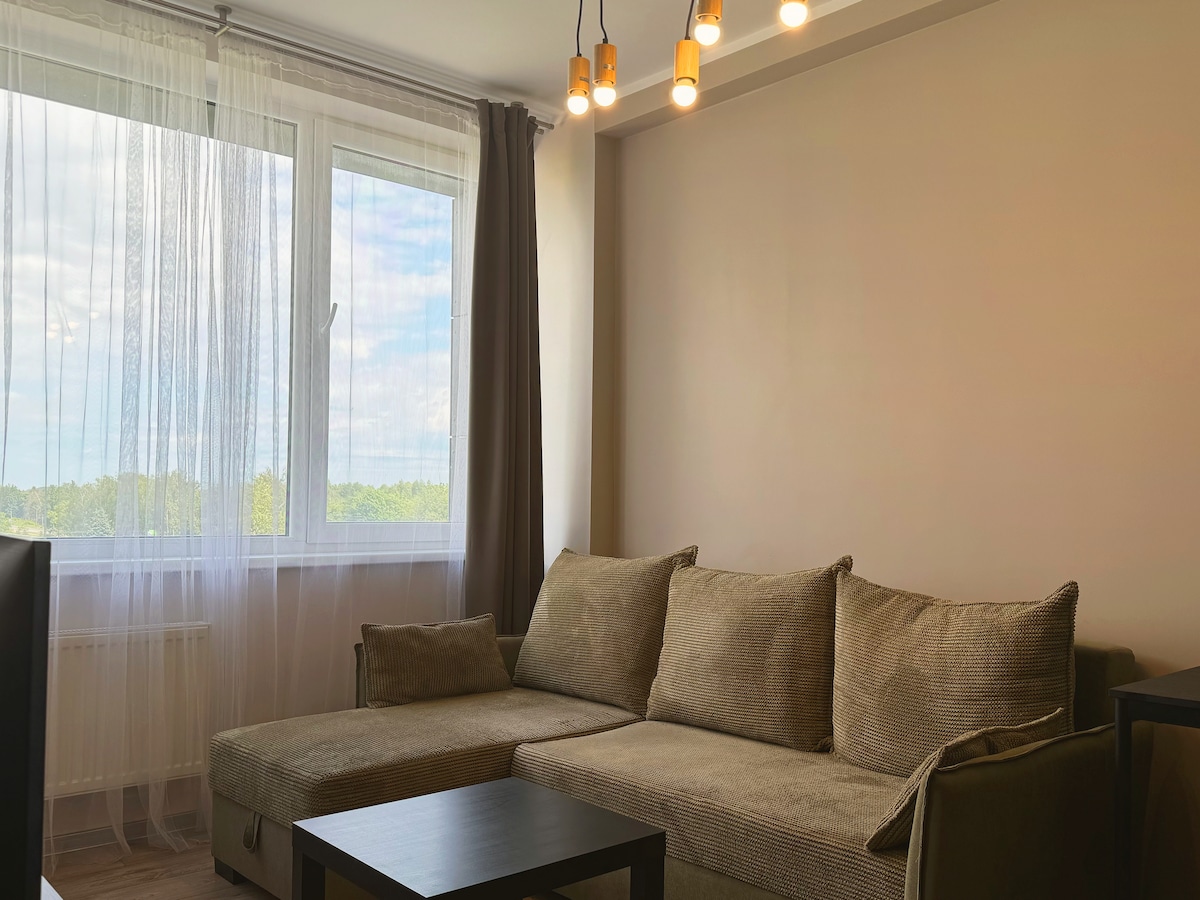A cozy living area features a comfortable light brown sectional sofa, complemented by a sleek black coffee table. Natural light filters through a large window, adorned with sheer curtains, allowing for a view of the greenery outside, while modern pendant lights illuminate the space.
