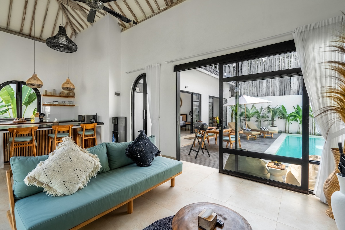 An open-plan living area features a comfortable turquoise sofa adorned with decorative pillows. Large glass doors provide a view of the pool area, which is surrounded by greenery. The dining area includes woven chairs and pendant lighting, contributing to a relaxed atmosphere.