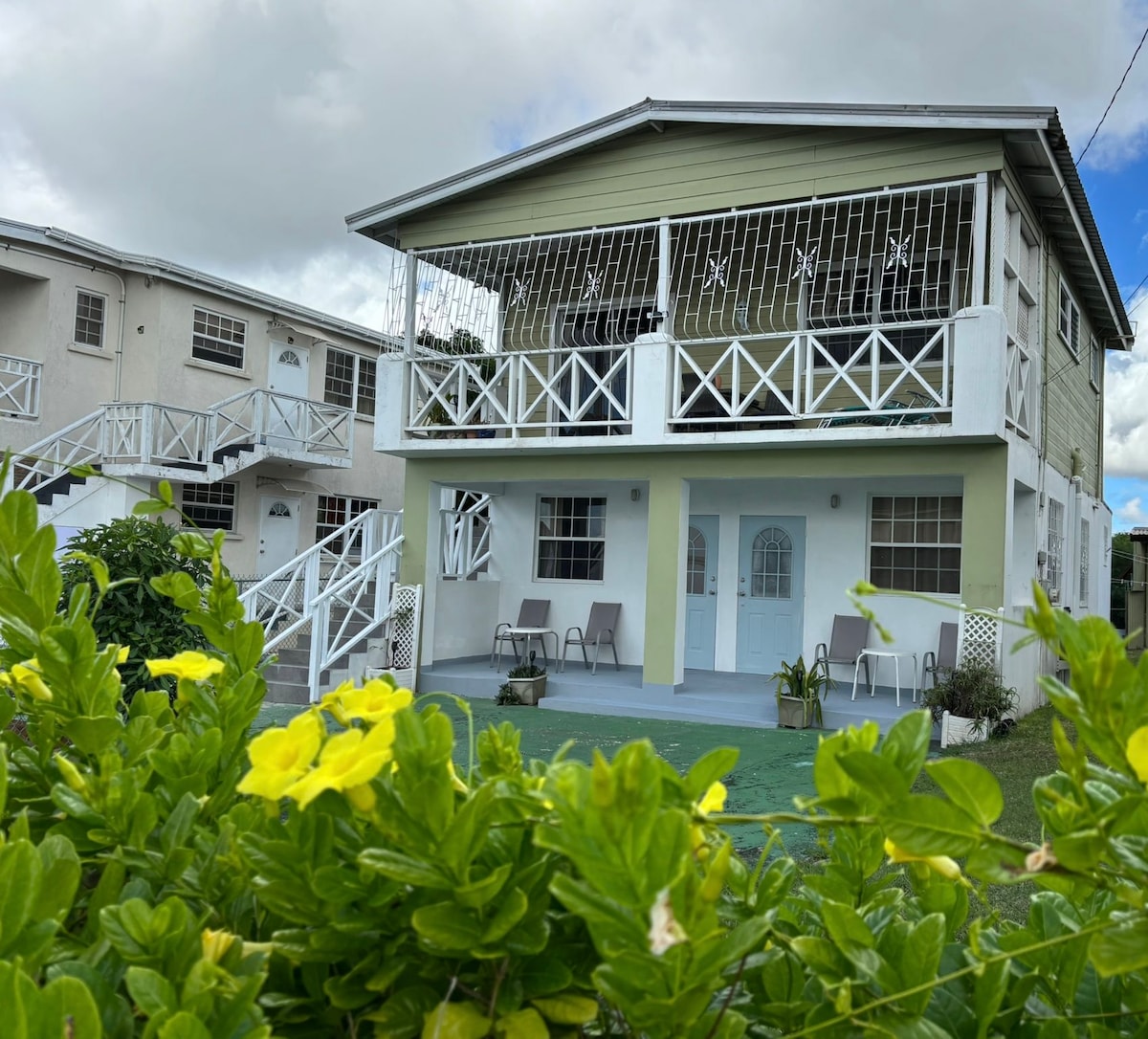 A two-story building is shown, featuring a spacious front patio with two chairs and a table. The structure has a light-colored exterior with large windows and decorative railings, surrounded by lush green foliage. The sky is partly cloudy, adding to the serene atmosphere.
