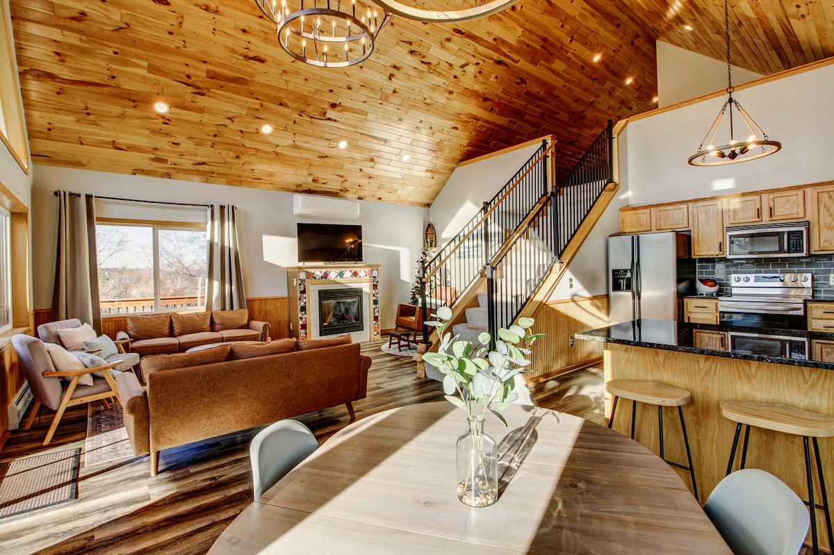 A spacious living and dining area is characterized by a cathedral ceiling with wooden beams. Natural light filters through large windows, illuminating a cozy seating arrangement and a dining table with chairs. The kitchen area features modern appliances and a breakfast bar, promoting a communal atmosphere.