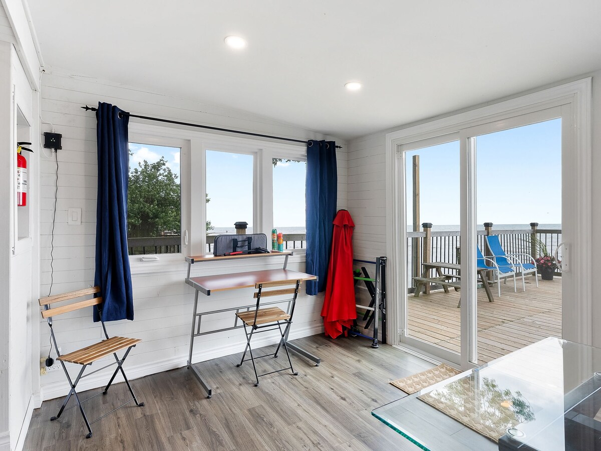 A bright interior space features large windows allowing natural light, with a small workspace setup including a table and two folding chairs. Soft blue curtains complement the scenic view of an outdoor deck, where additional seating is visible.