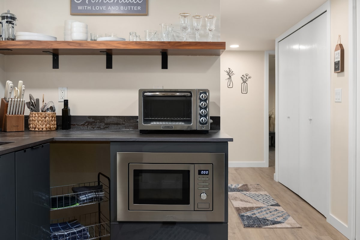 A modern kitchen area is featured, showcasing sleek dark cabinetry and open shelving. Kitchen appliances include a compact oven and a microwave, while utensils and glassware are organized neatly. The space is complemented by a decorative wall sign and a patterned rug on the floor.