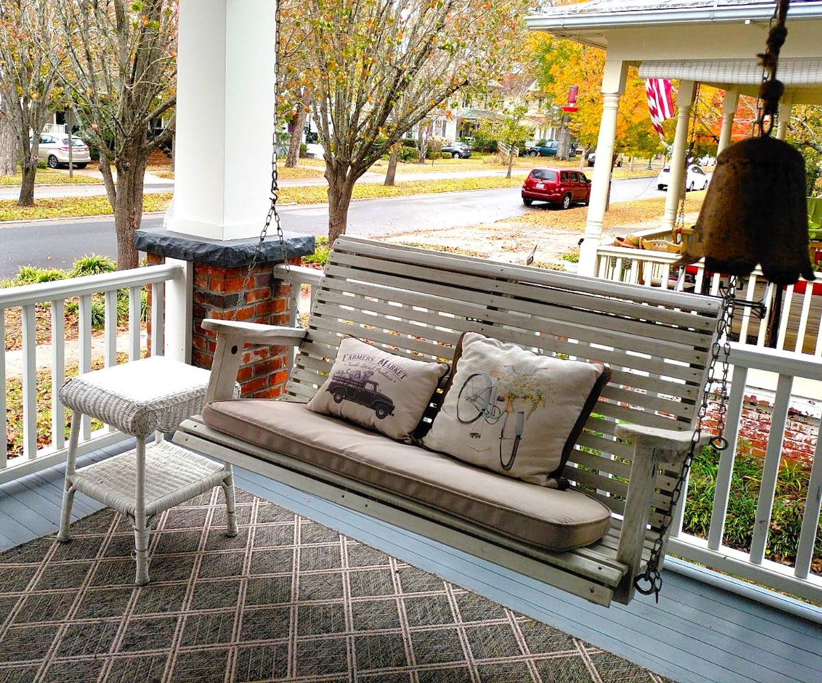 A spacious front porch features a white swing with a cushion and decorative pillows. A small side table sits beside the swing, contributing to the cozy seating area. Trees line the street, showcasing seasonal foliage and inviting a peaceful atmosphere.