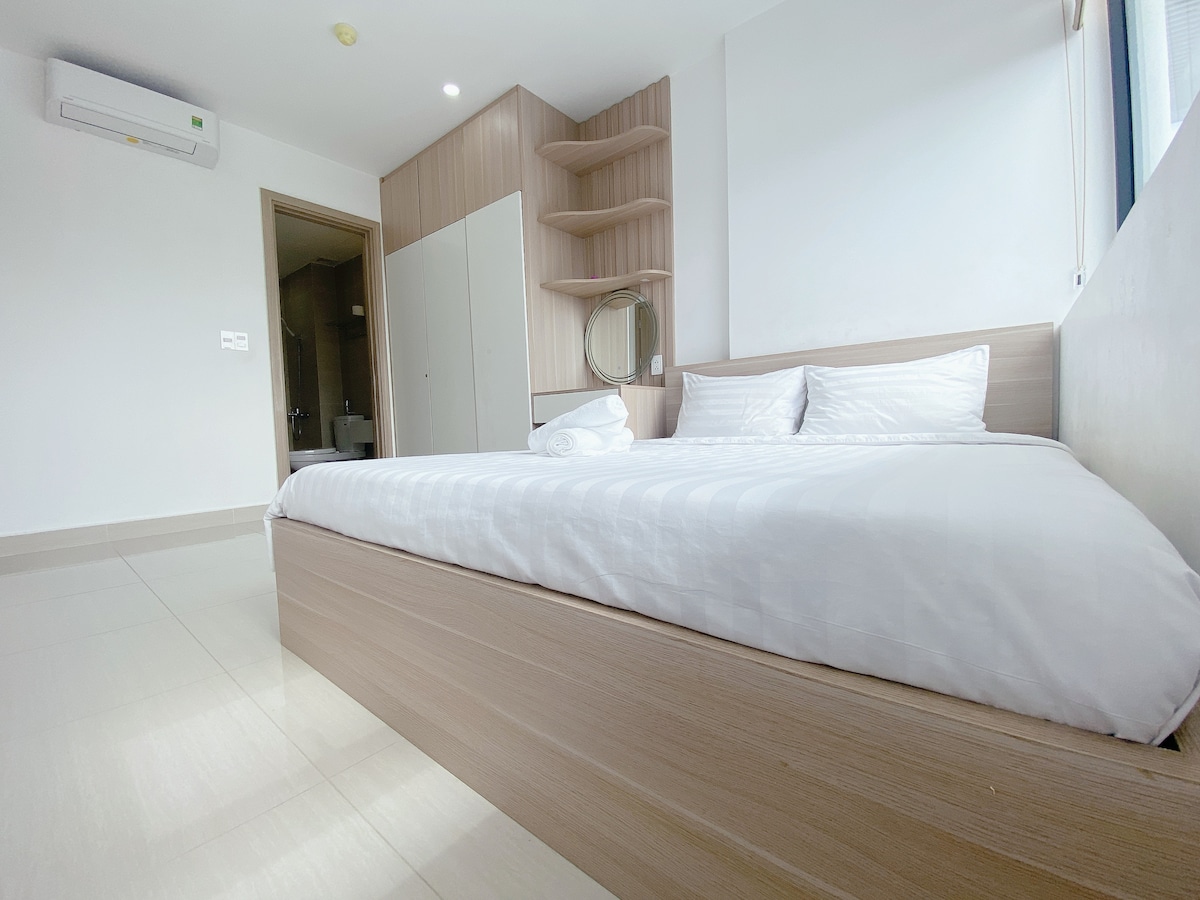 A spacious bedroom features a light wooden bed with neatly arranged white bedding. Soft natural light enters through a large window, illuminating the room's minimalist decor. A mirror is mounted on the wall above a small shelf, and a doorway leads to an adjoining bathroom.