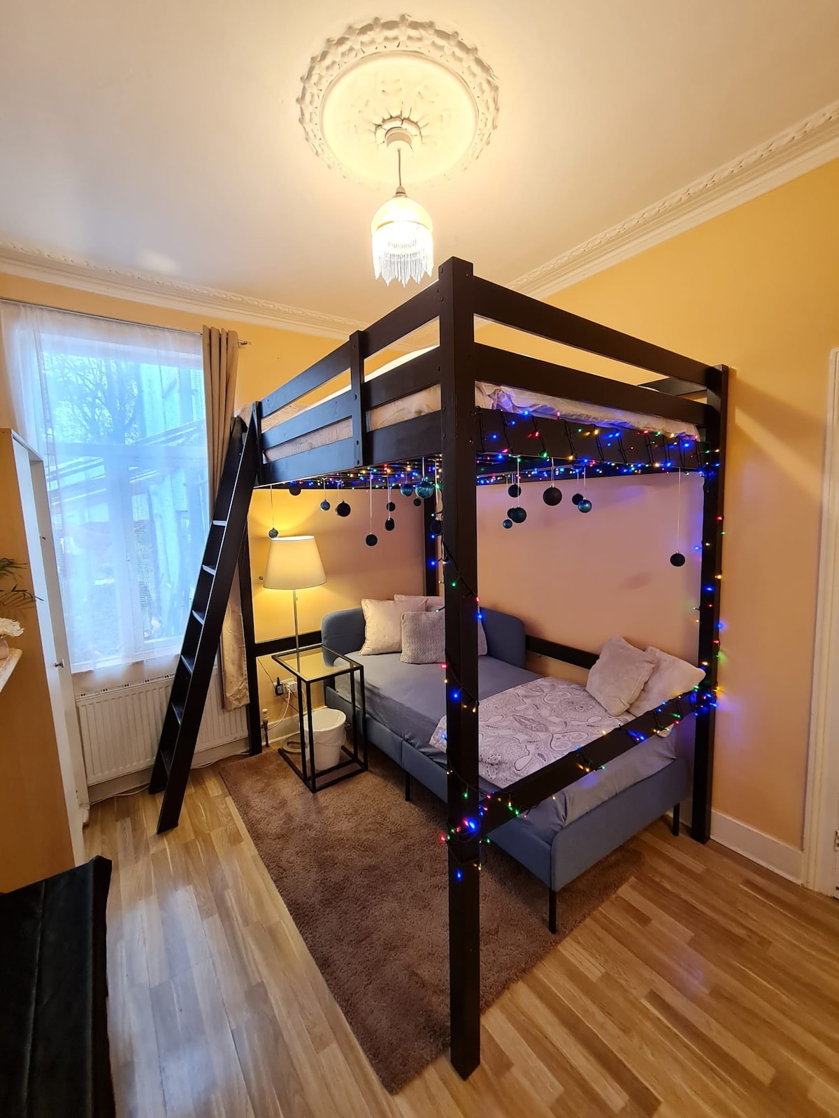 A lofted bed frame with a cozy sleeping area underneath is adorned with colorful lights. Soft bedding creates an inviting space, while a climbing ladder provides access to the elevated bed. A lamp beside the bed adds to the room's ambiance, with natural light filtering in through the window.