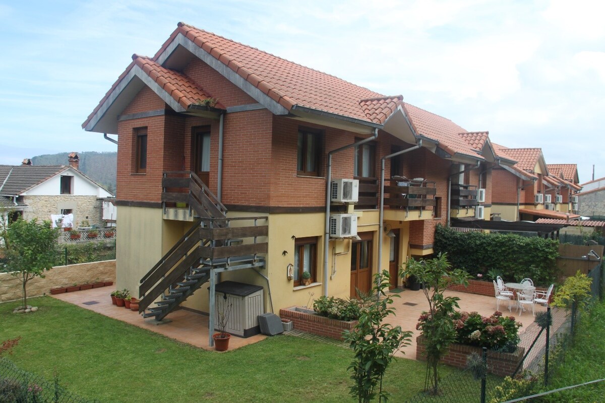 The exterior of a welcoming apartment building is shown, featuring a two-story structure with a red-tiled roof and balconies. Air conditioning units are visible on the side. Surrounding greenery includes trees and well-maintained grass, creating a peaceful environment.